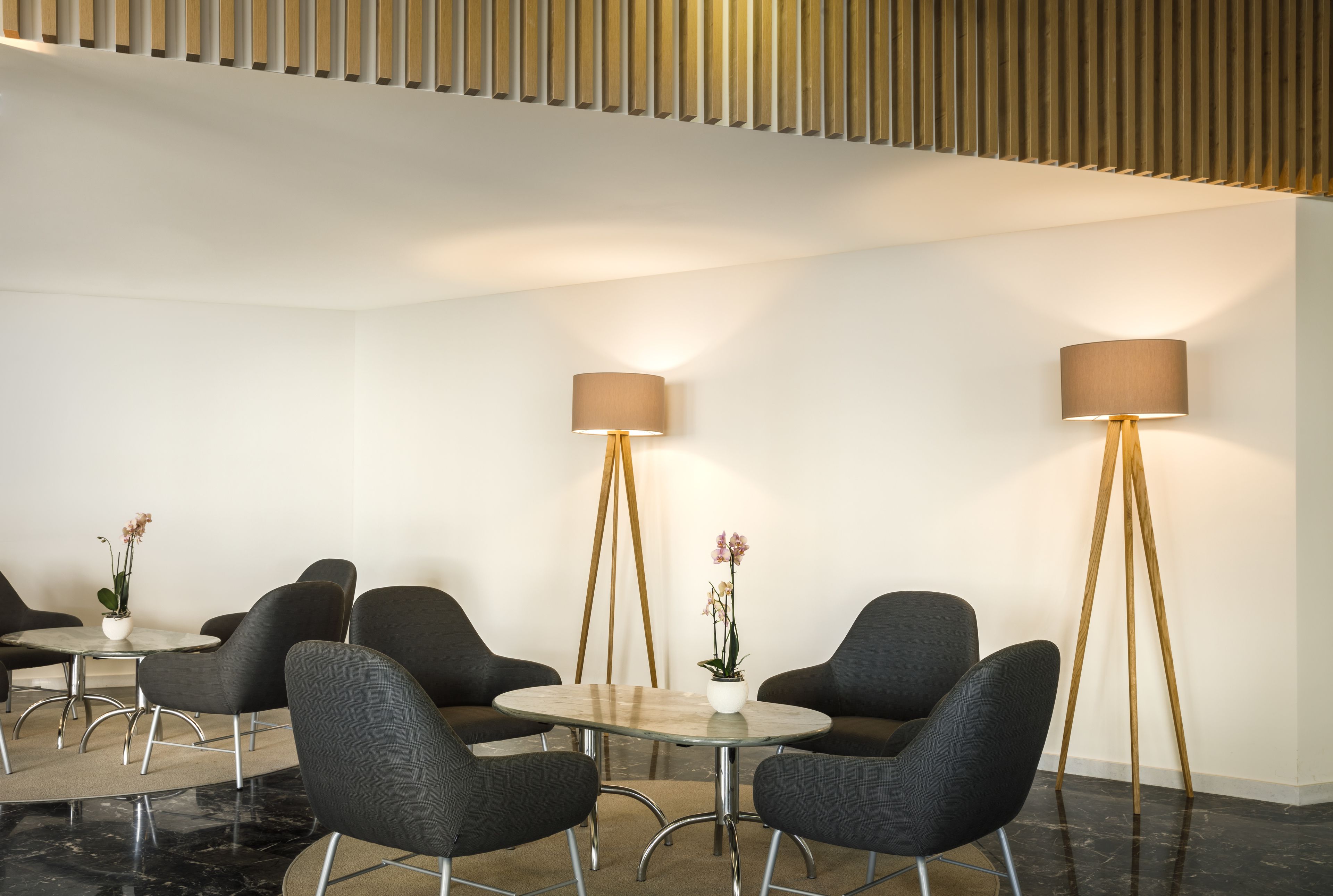 A modern waiting area with a round table, four black chairs, two floor lamps, and potted plants.