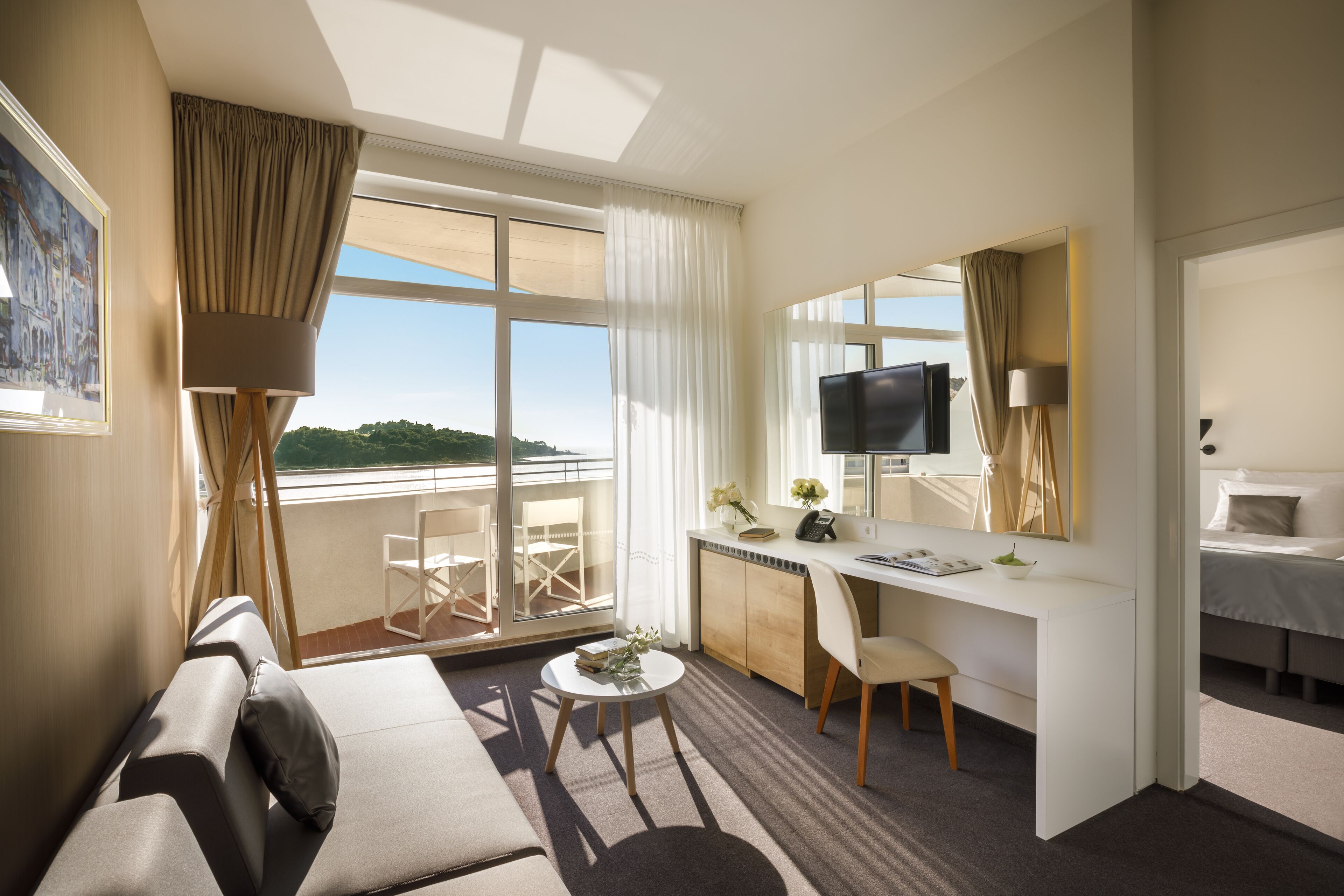 A hotel room with a sofa, a desk, a TV, and a balcony with a view of the greenery.