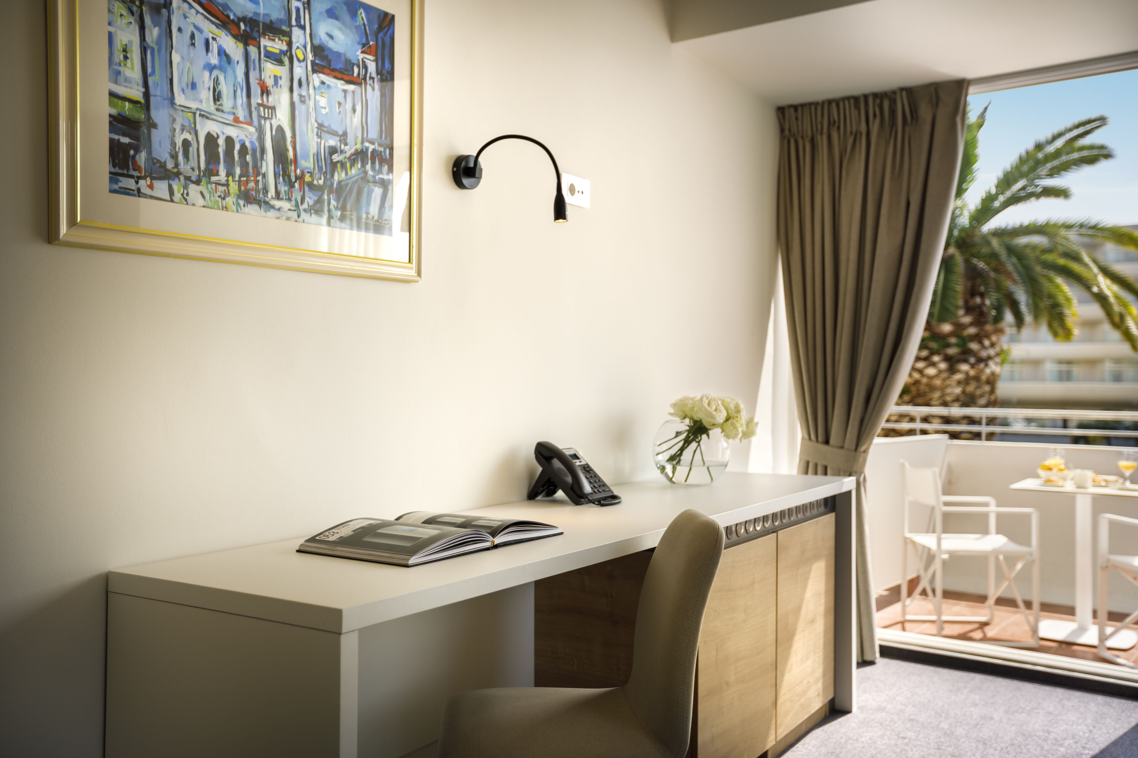 A hotel room with a desk, a telephone, a vase with flowers, and a view of the outdoors.