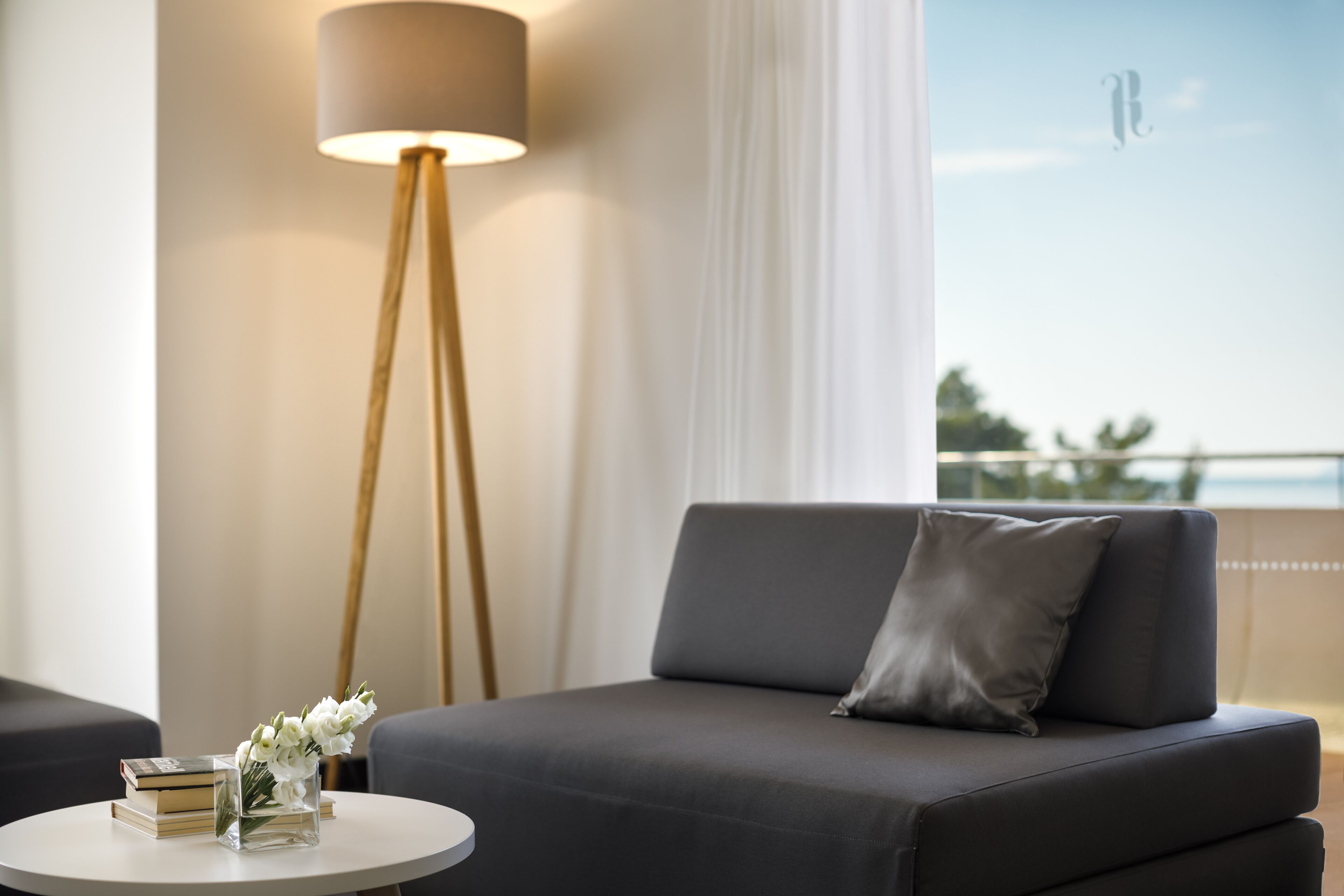 A modern living room with a sofa, a side table with books and flowers, a floor lamp, and a view of the outdoors.