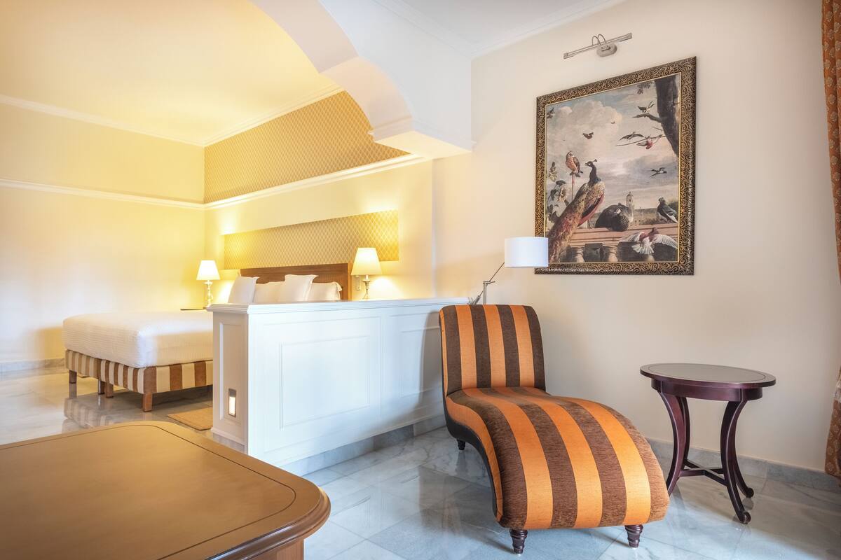 A hotel room with a bed, a striped armchair, a small table, and a painting on the wall.