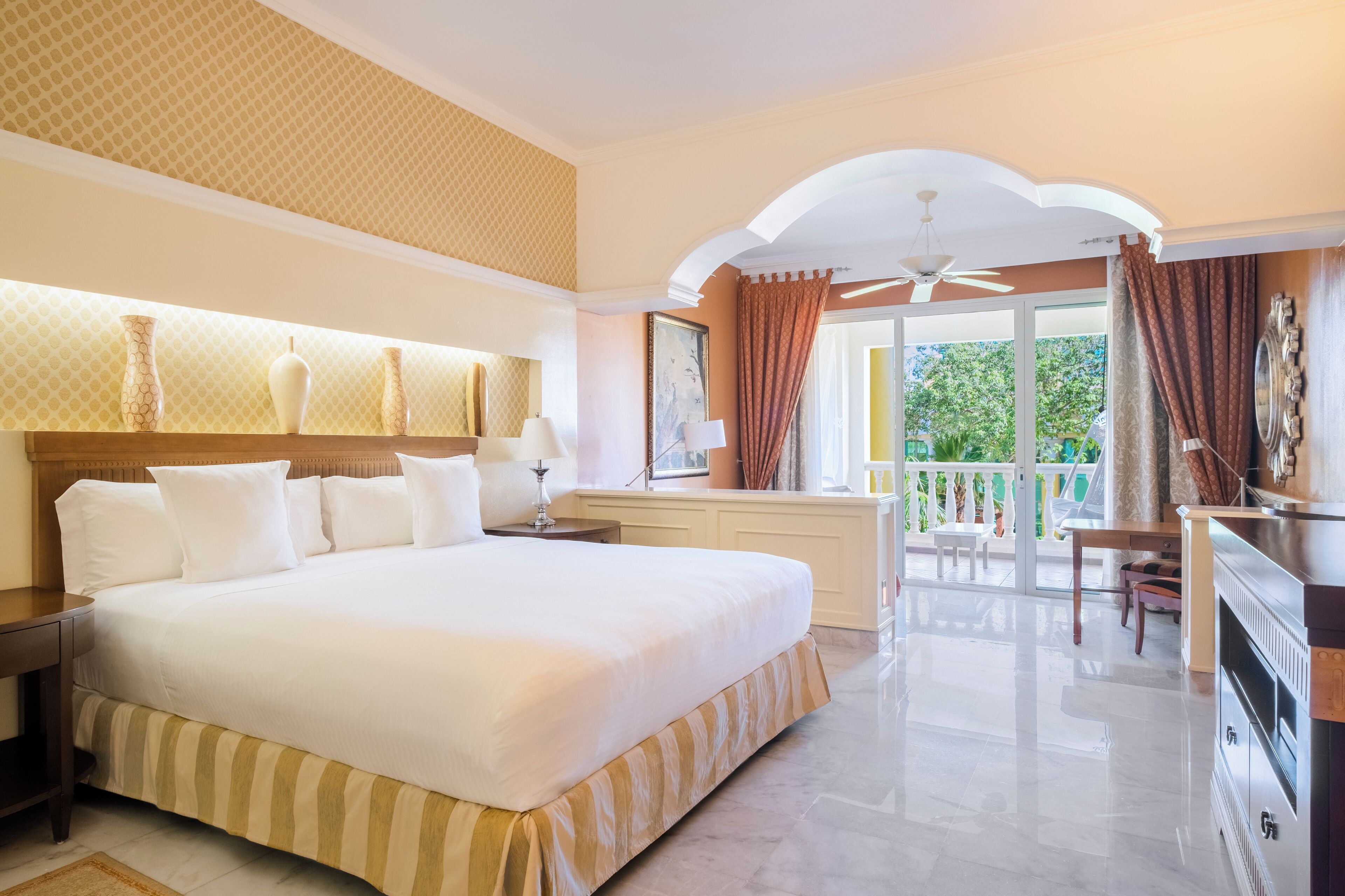 A hotel room with a large bed, a desk, a chair, a ceiling fan, and a balcony with a view of greenery.