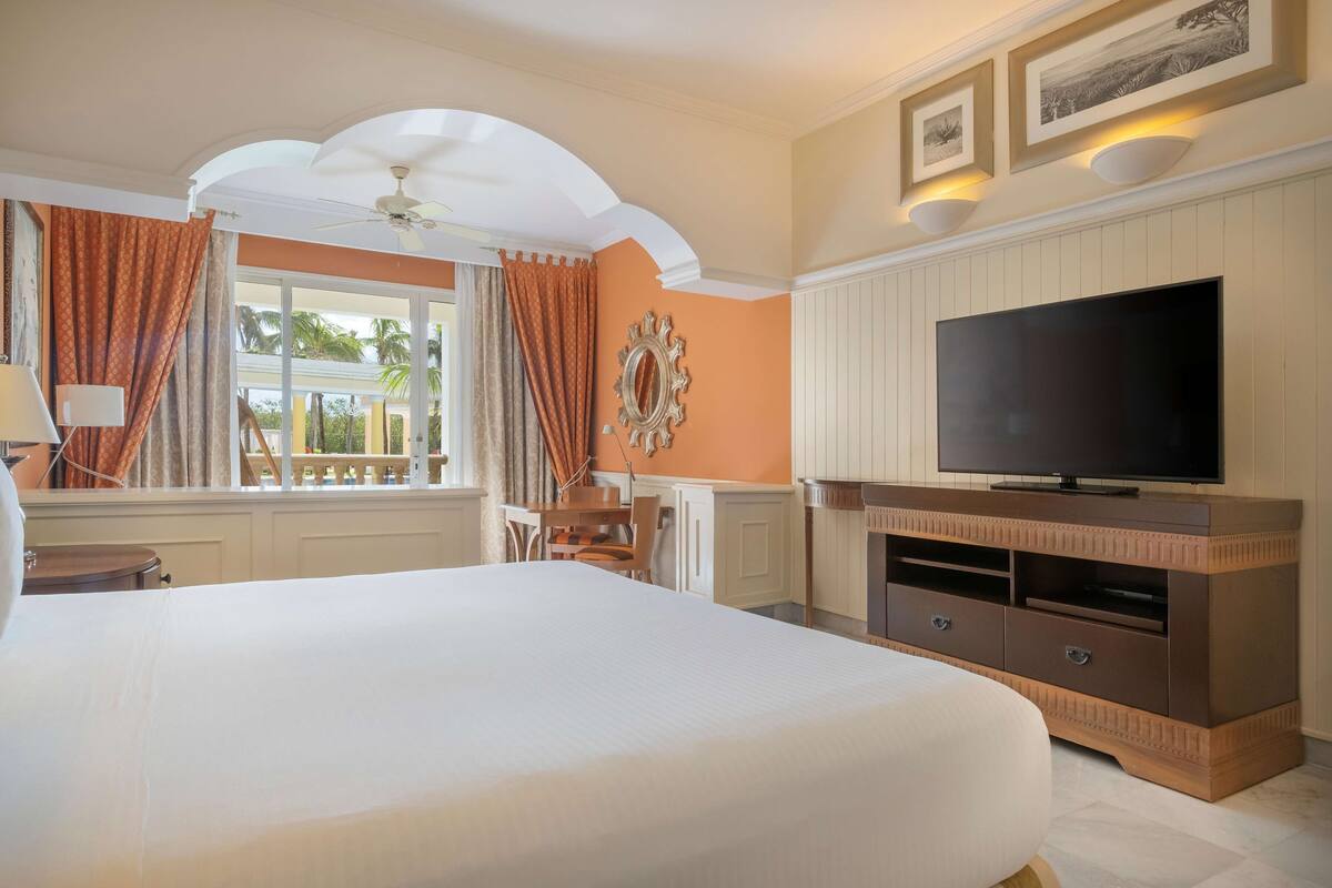A hotel room with a large bed, a television, a dining table, and a view of palm trees outside.