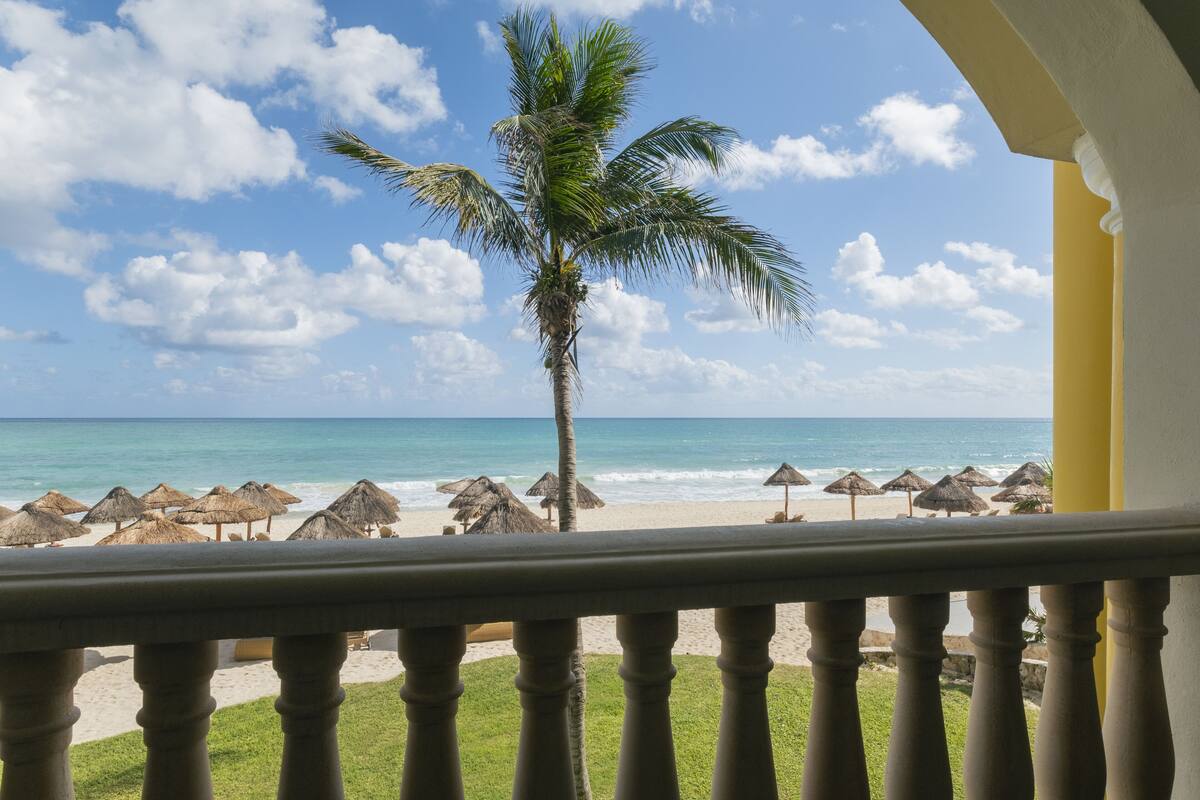 A balcony with a view of a beach, palm trees, and thatched umbrellas.