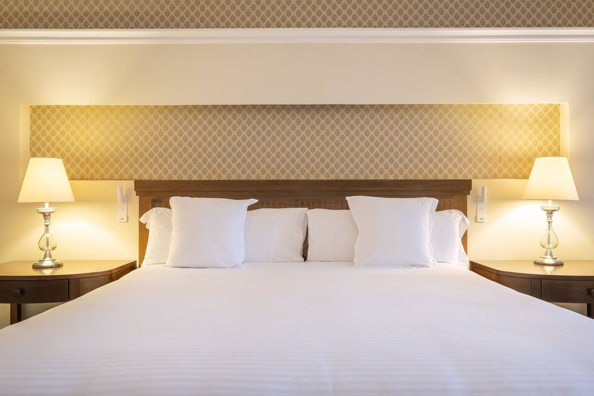 A neatly made bed with white linens and pillows, flanked by two bedside tables with lamps, set against a patterned wallpaper backdrop.