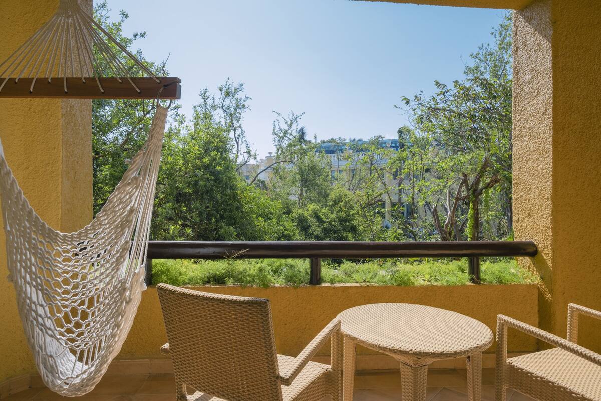 A balcony with wicker furniture, a hanging hammock, and a view of greenery and buildings.