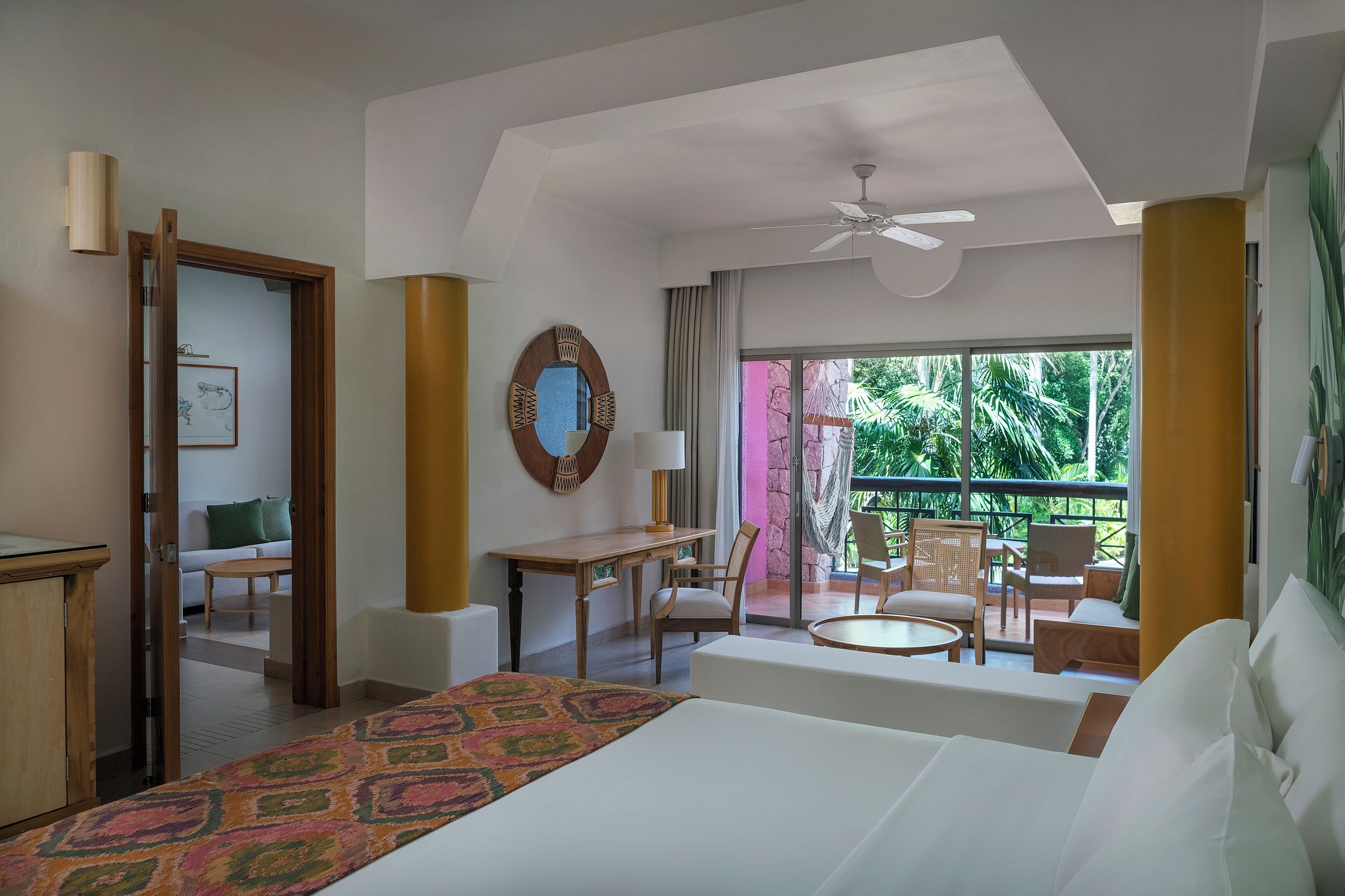 A hotel room with a bed, a desk, a chair, a balcony with outdoor seating, and a ceiling fan.