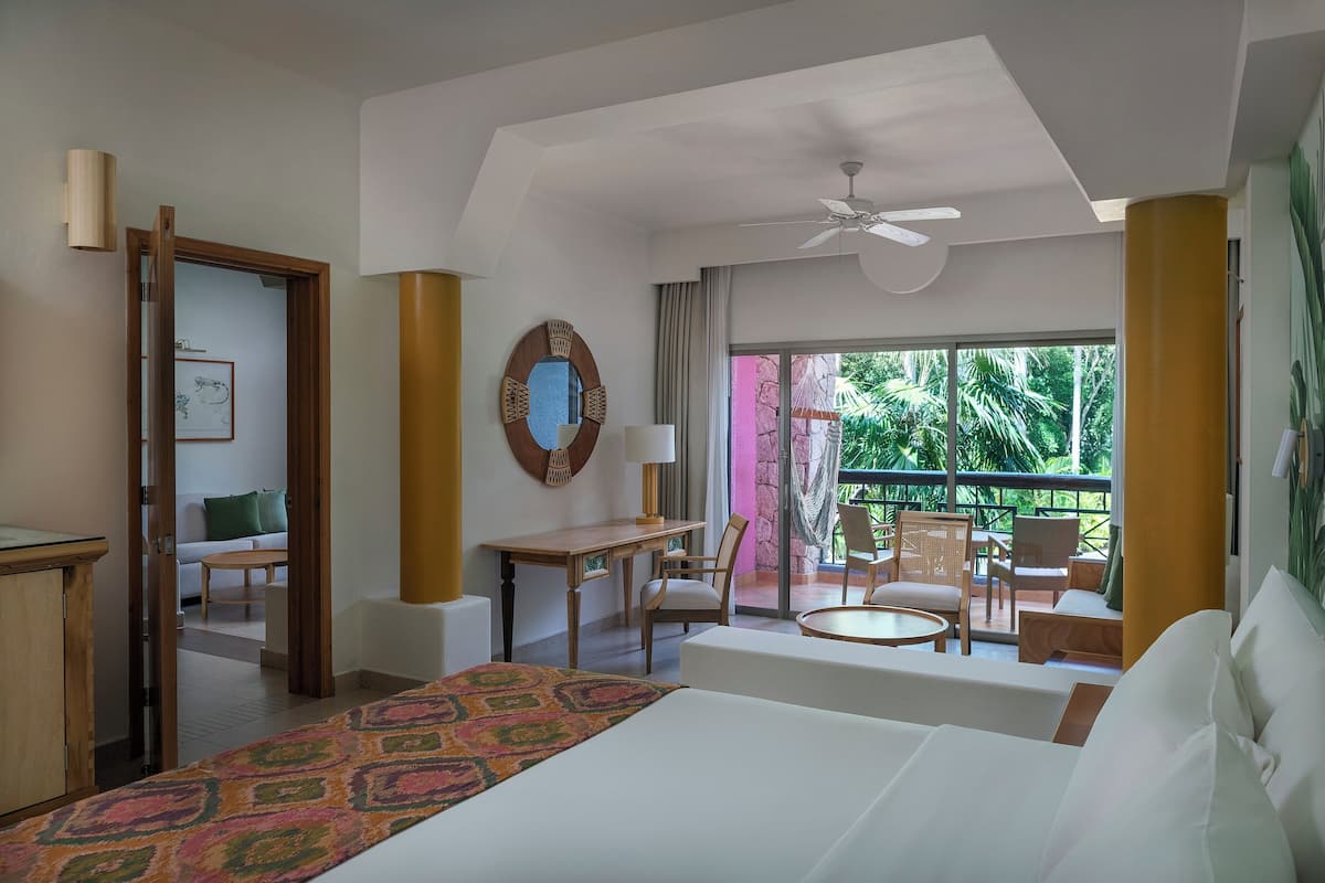 Habitación de hotel con cama, escritorio, silla, balcón con mobiliario exterior y ventilador de techo.