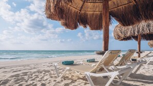 Ein Strand mit weißen Liegestühlen und einem Reetdach-Sonnenschirm, feiner Sand und das Meer im Hintergrund.