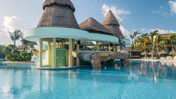 Ein Poolbereich eines Resorts mit strohgedeckten Gebäuden, Palmen und einem klaren, blauen Himmel.