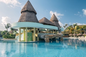 A resort pool area with thatched-roof structures, palm trees, and a clear blue sky.