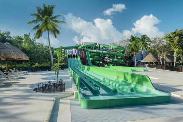 A water park with a green water slide, palm trees, and thatched-roof huts.