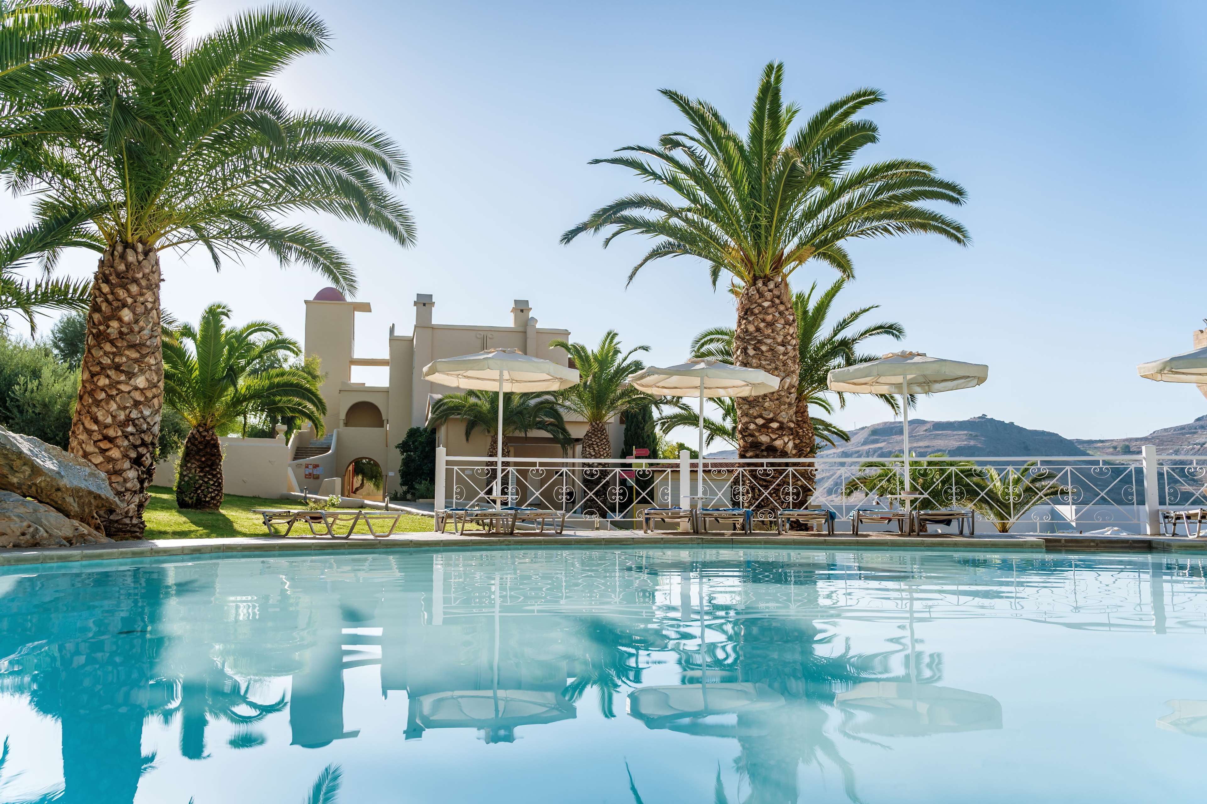 A swimming pool with lounge chairs and umbrellas, surrounded by palm trees and a building with arches.