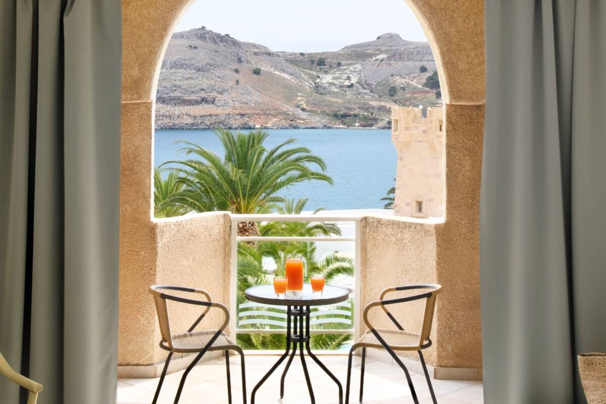 A balcony with a view of the sea and hills, two chairs, a table with drinks, and a woven bag.