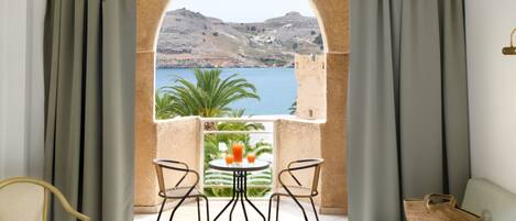 A balcony with a view of the sea and hills, two chairs, a table with drinks, and a woven bag.