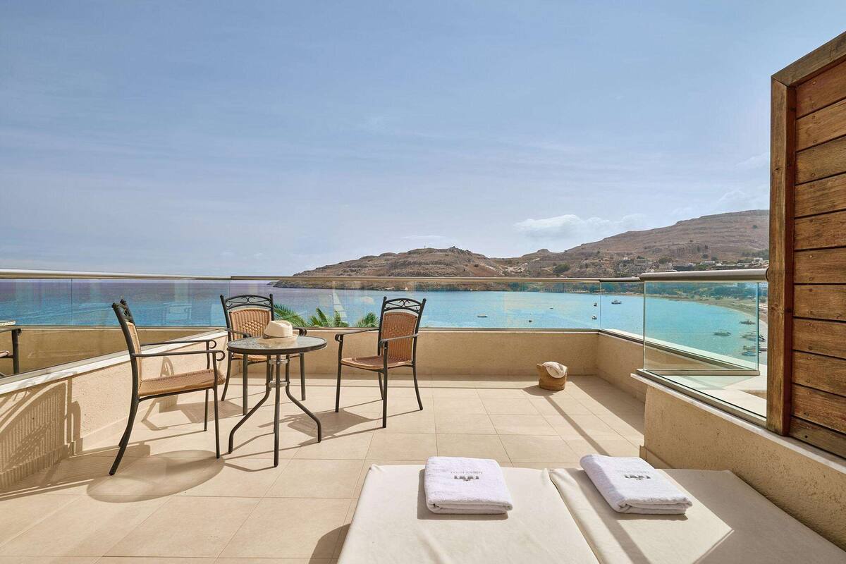 A balcony with a view of the sea, two wicker chairs, a small round table, and two towels on a lounge chair.