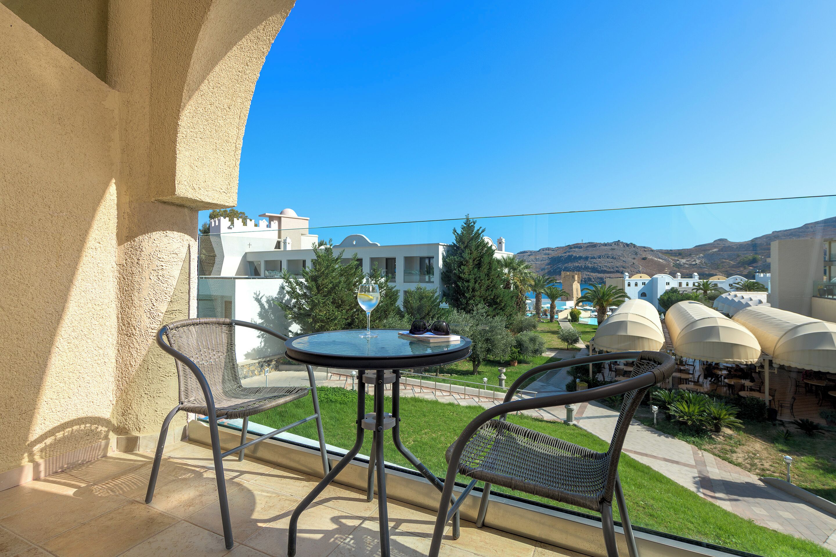 A balcony with two wicker chairs and a small round table, overlooking a pool area with umbrellas and a view of buildings and hills.