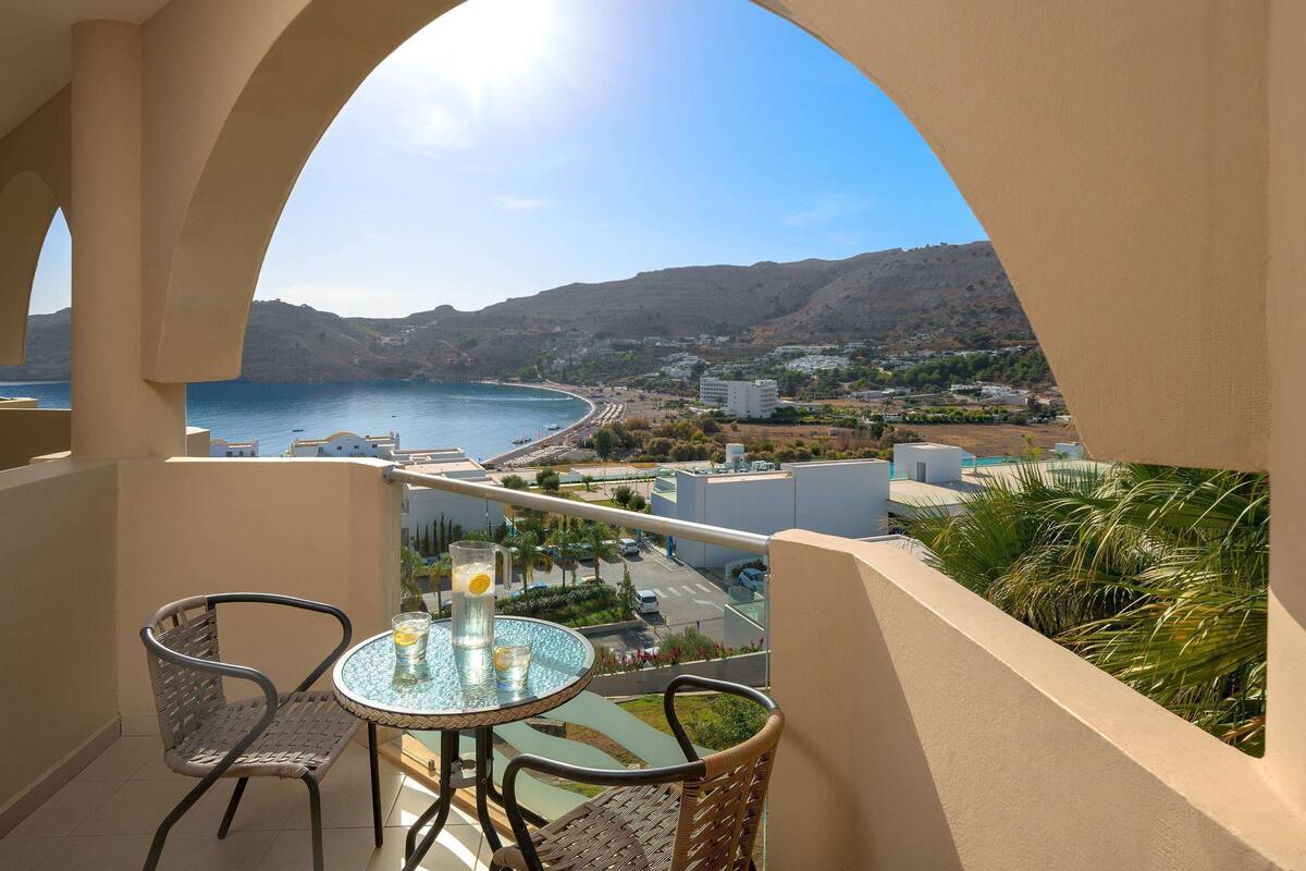 A balcony with a table and chairs, overlooking a coastal town and the sea.