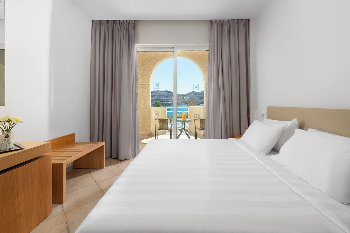 A hotel room with a large bed, a wooden desk, and a view of a balcony with a table and chairs.