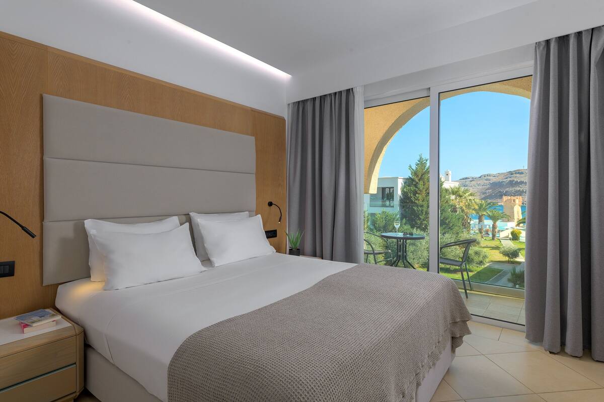 A hotel room with a large bed, a nightstand, and a view of a balcony with a table and chairs.