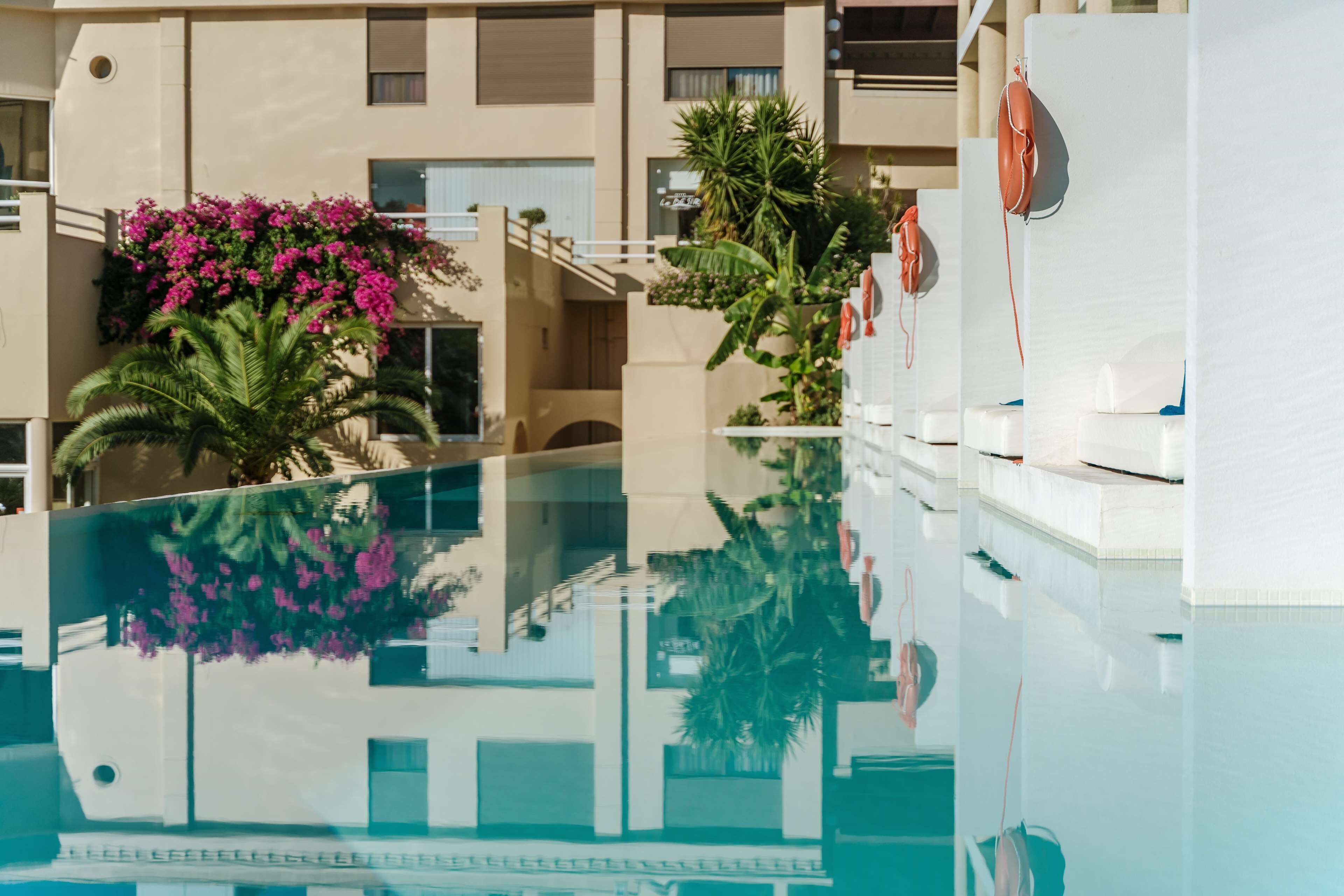 Zona de piscina moderna de hotel con paredes de vidrio transparente, una palmera y flores rosas.