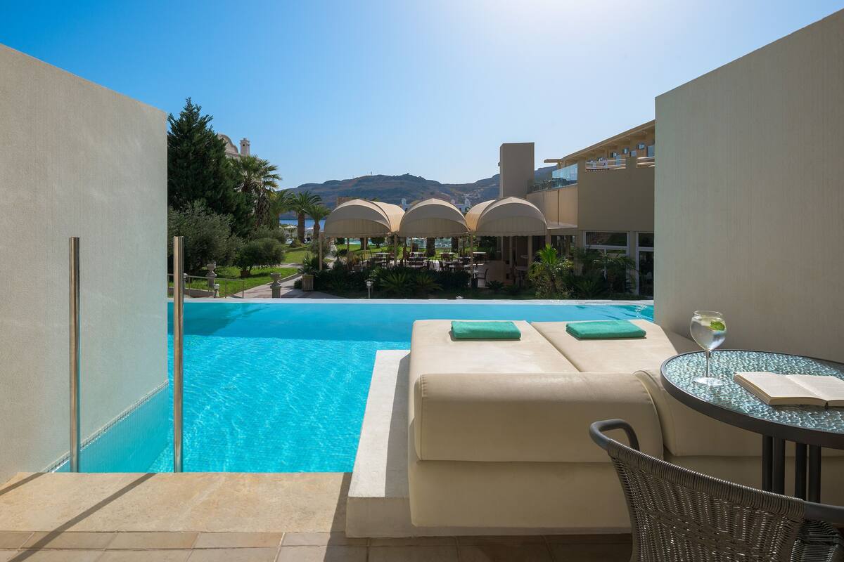 A poolside area with a lounge chair, a glass table with a book and a glass of water, and a view of the pool and distant mountains.