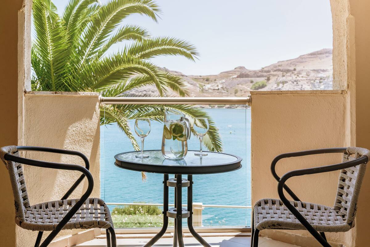 A balcony with two wicker chairs, a small round table, and a glass pitcher with glasses on it, overlooking a body of water and palm trees.