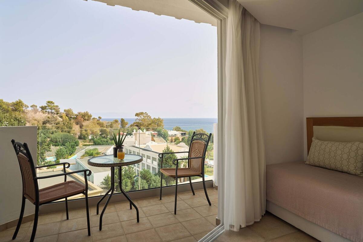 A balcony with a table and chairs, a bed, and a view of the sea and buildings.