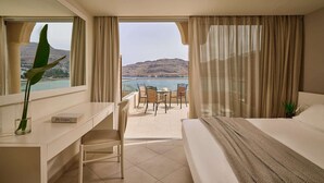 A hotel room with a bed, a desk, a chair, and a view of the sea and mountains.