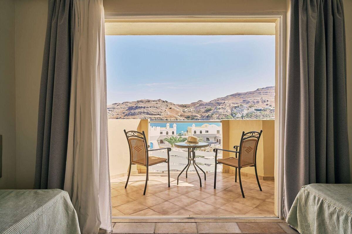 A balcony with two wicker chairs and a small round table, overlooking a coastal town and hills.
