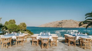 A dining area with tables and chairs set up for outdoor dining, overlooking a body of water and a mountainous landscape.