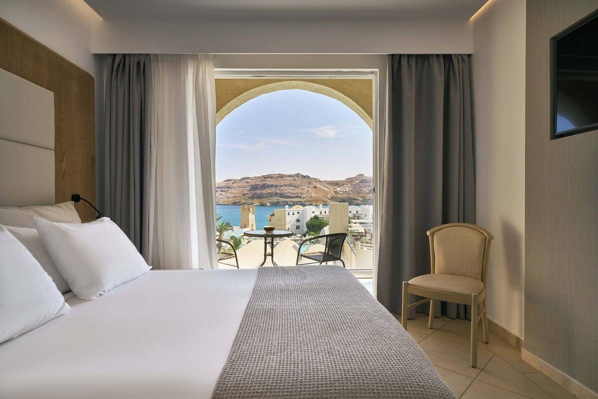 A hotel room with a large bed, a small table, a chair, and a view of the sea and hills.