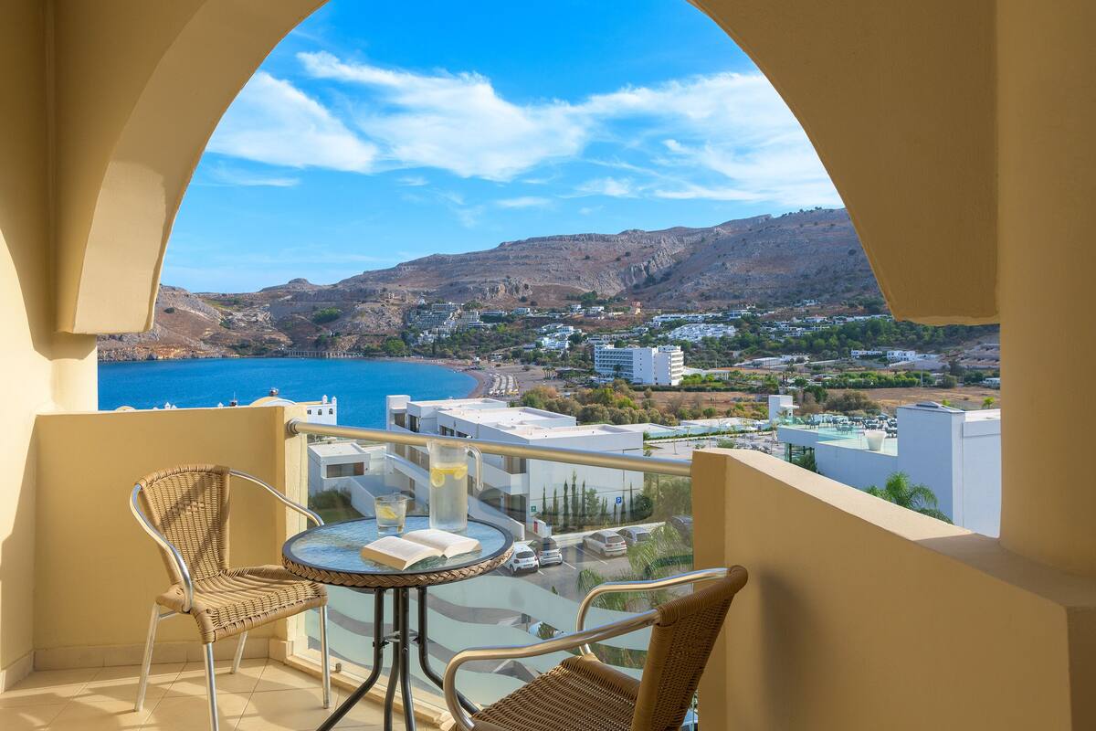 A balcony with wicker chairs, a small table, and a clear view of a coastal town and mountains.
