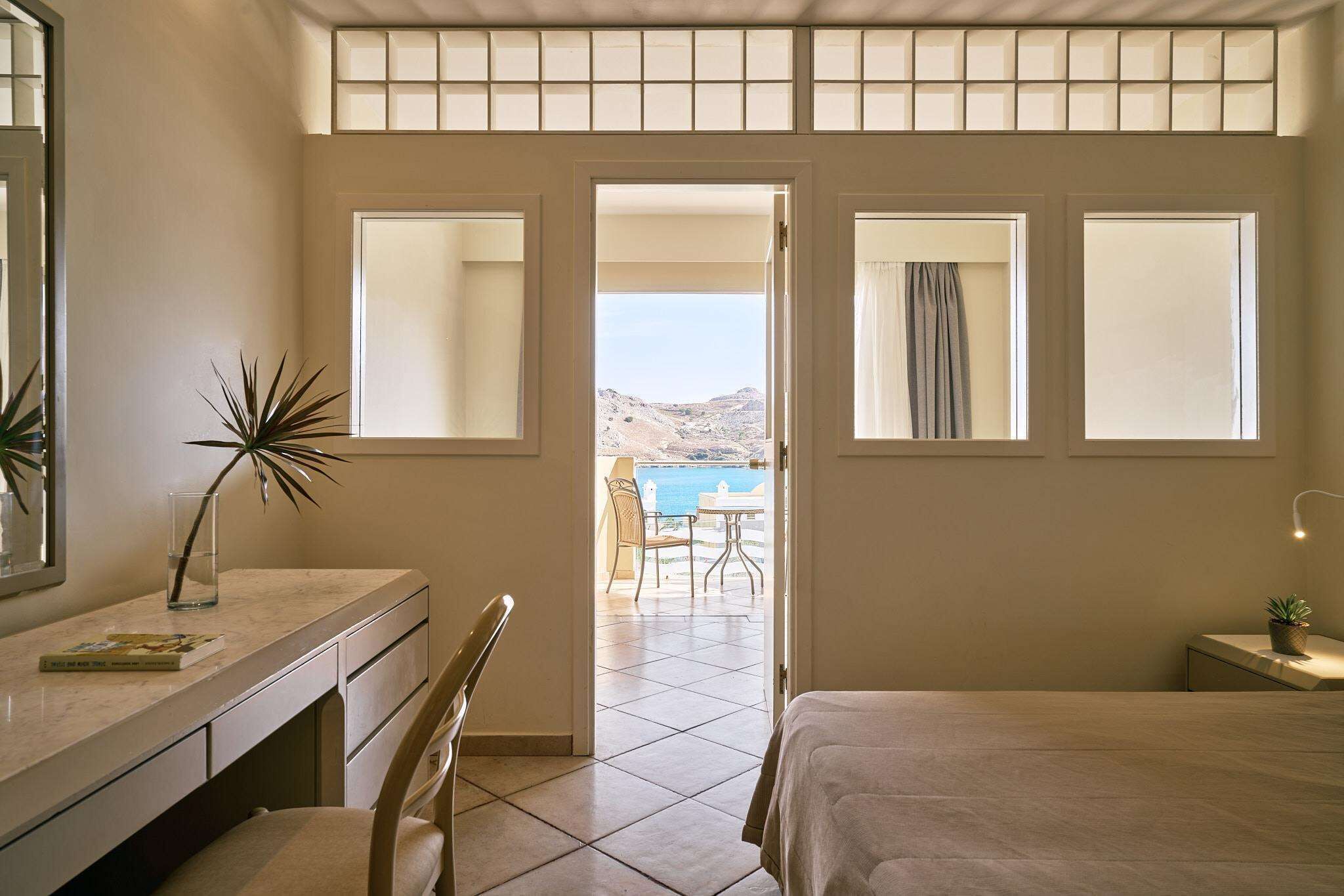 Un dormitorio con cama, escritorio, silla y vista al mar a través de una puerta.