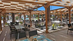 An outdoor patio area with tables and chairs, a pool in the background, and palm trees.