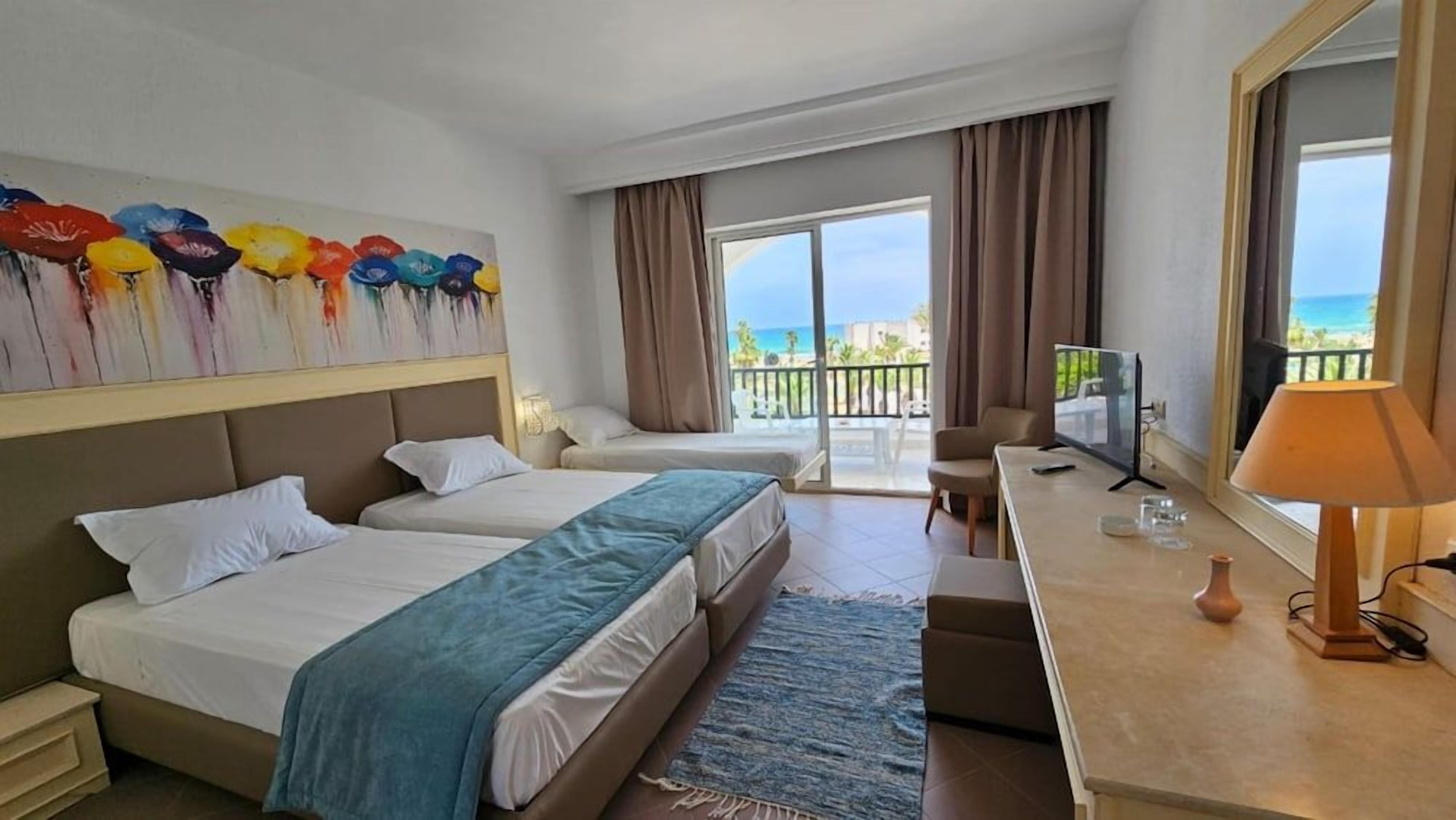 A hotel room with two beds, a desk, a chair, a TV, and a balcony with a view of the sea.