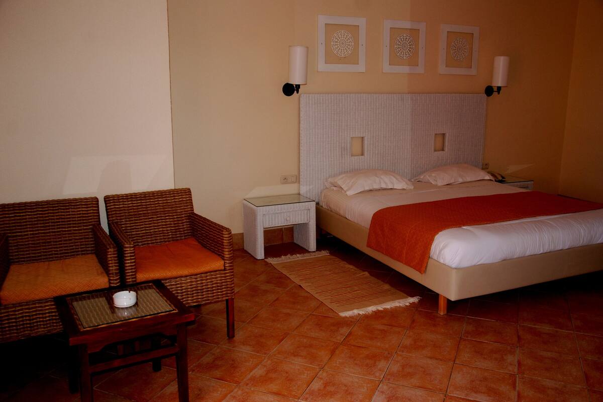 A hotel room with a bed, two chairs, a nightstand, and a wall-mounted lamp.