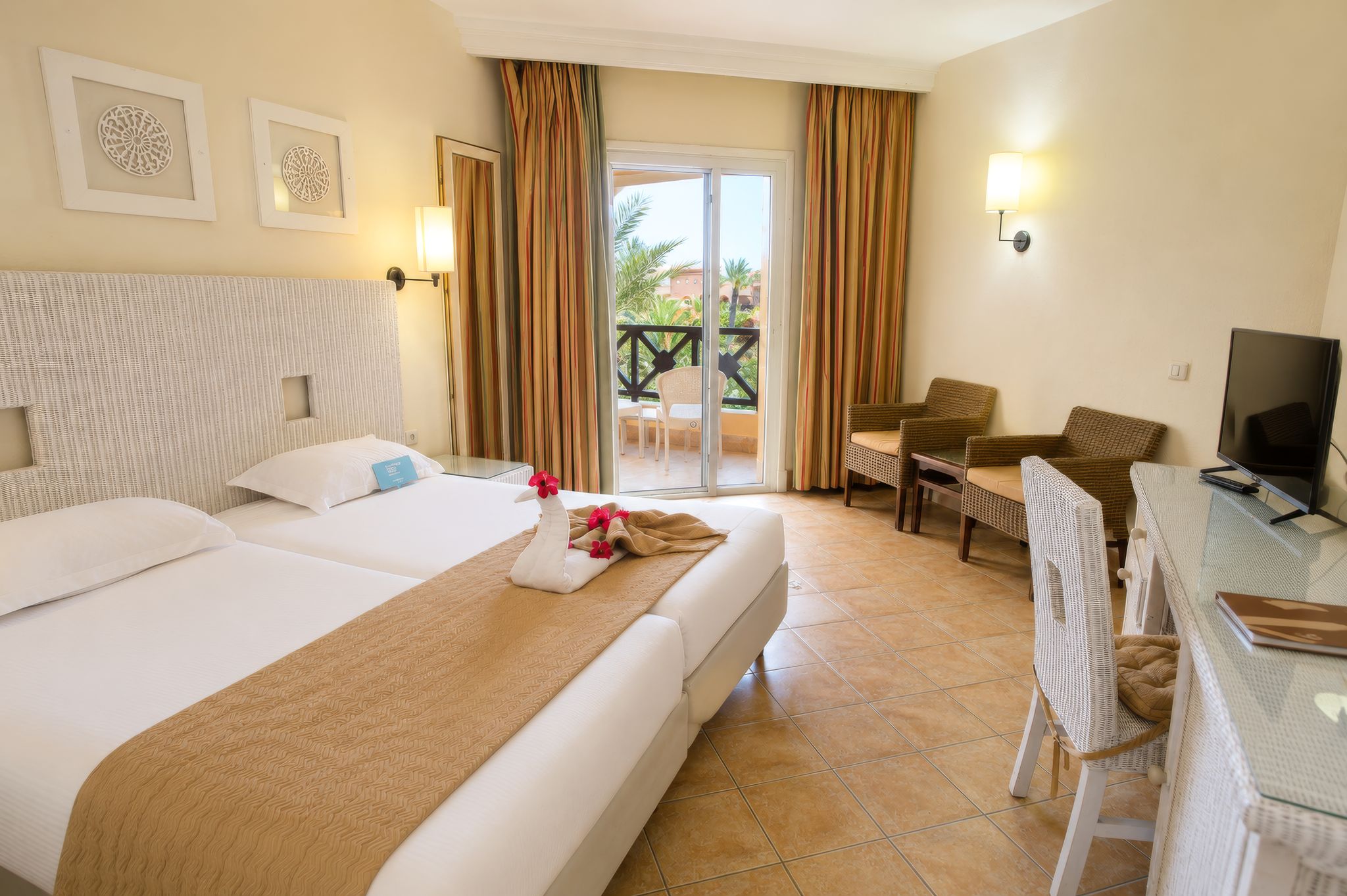 A hotel room with two beds, a balcony with curtains, a television, and a dining area.