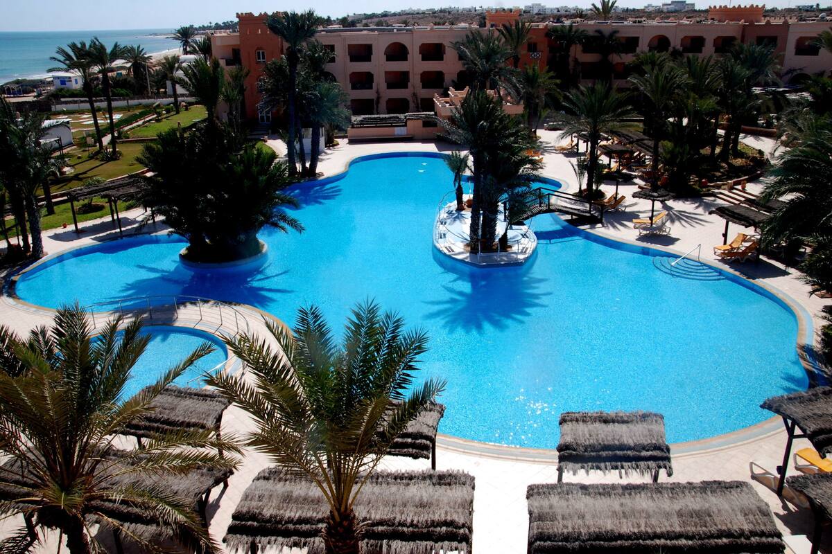 A large, clear blue pool surrounded by palm trees and lounge chairs, with a building in the background.