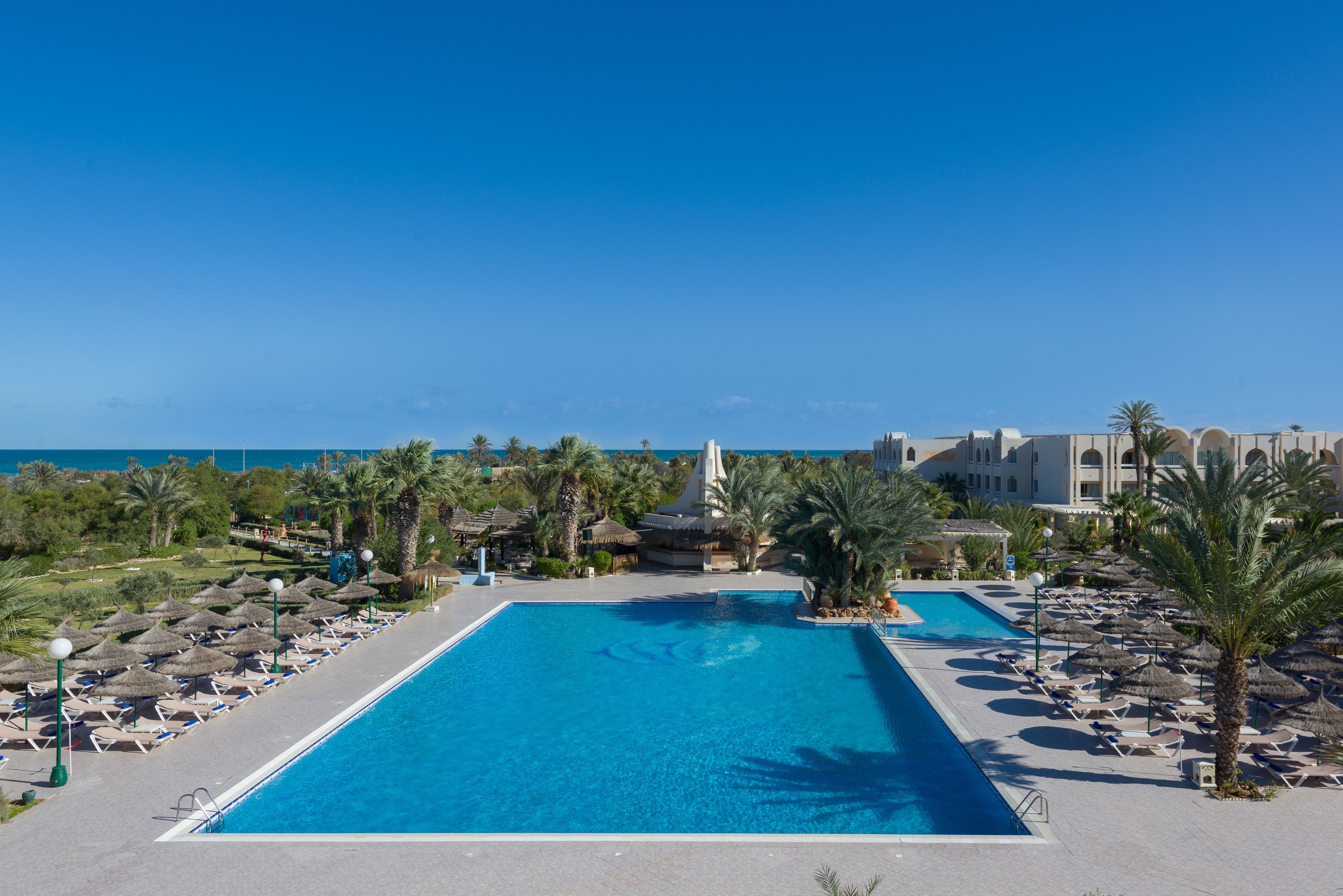 A resort pool with lounge chairs and umbrellas, surrounded by palm trees and buildings.