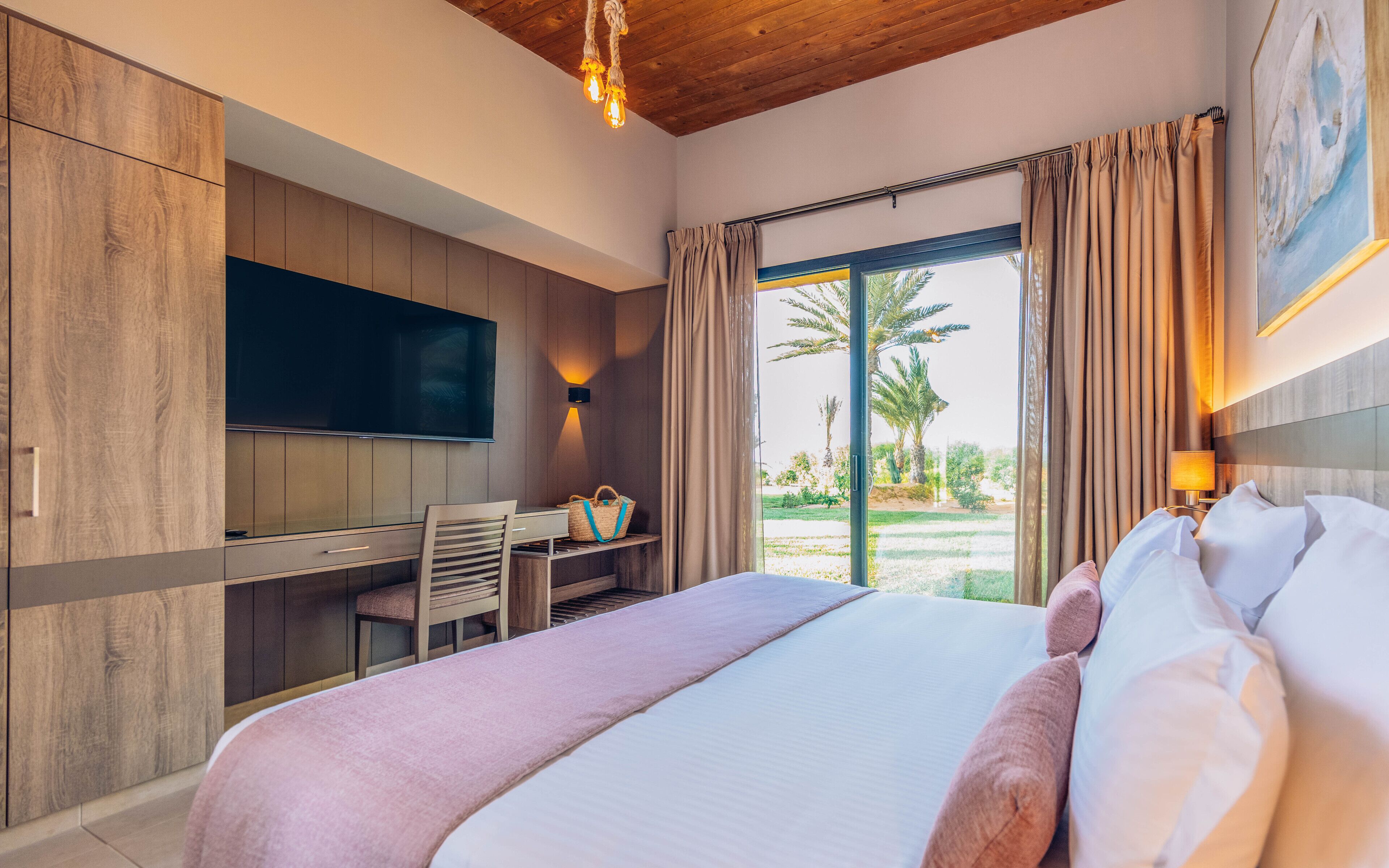 A hotel room with a bed, a desk, a television, and a view of palm trees.