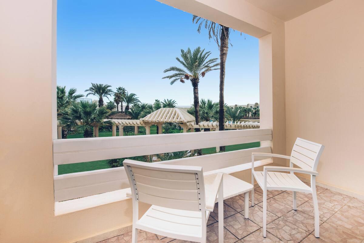 A balcony with white chairs and a table, overlooking a garden with palm trees and a gazebo.