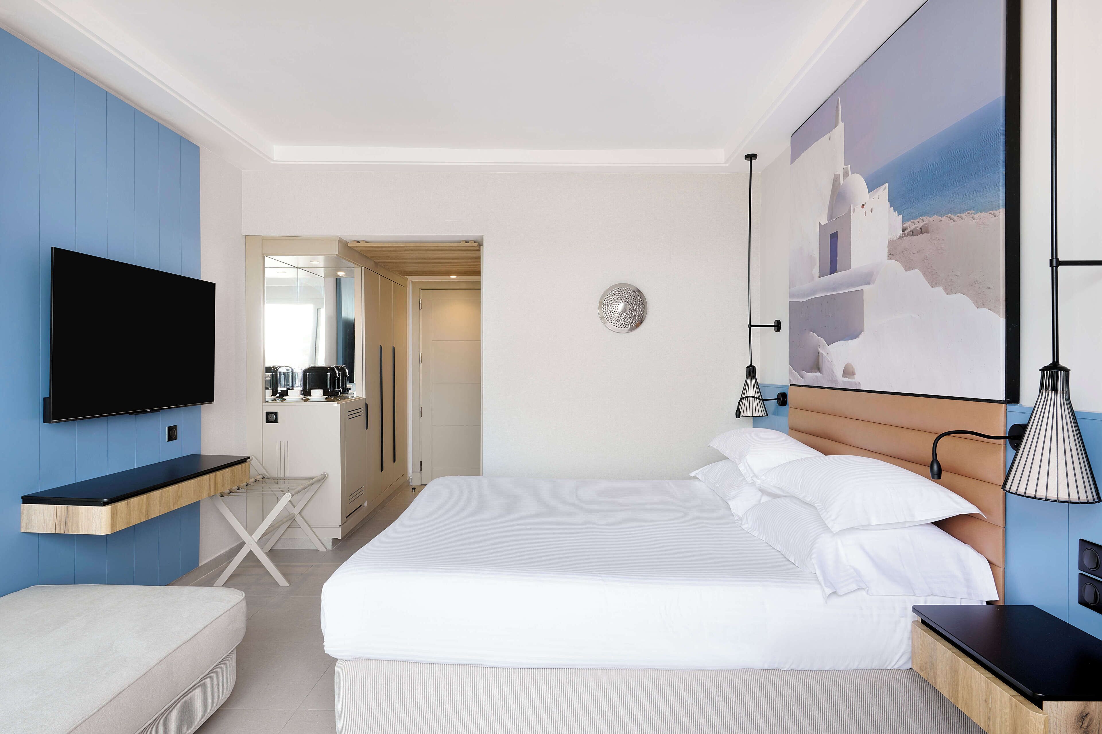 A hotel room with a large bed, a flat-screen TV mounted on the wall, a small kitchenette, and a painting of a coastal scene.