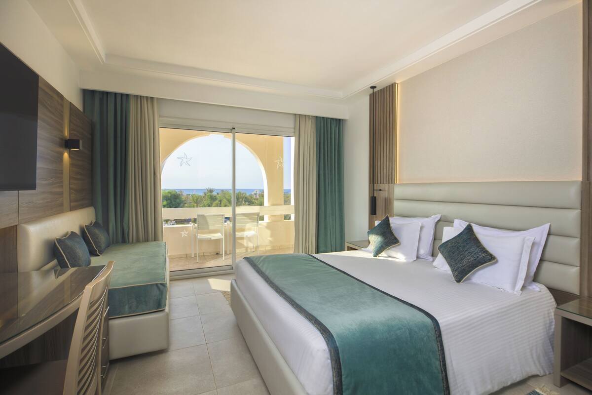A hotel room with a large bed, a seating area with a table and chairs, a TV, and a balcony with a view of the sea.
