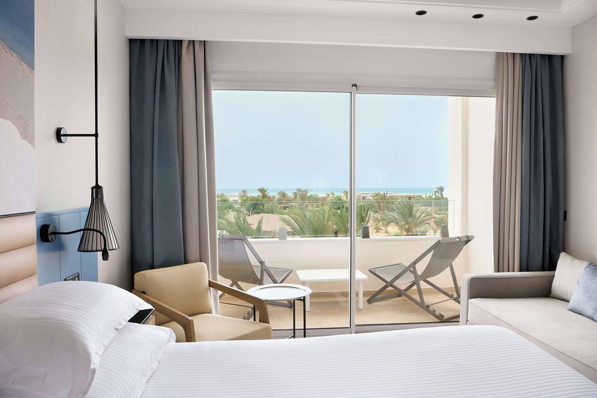 A hotel room with a bed, two chairs, a small table, and a balcony with a view of the beach and palm trees.