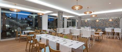 Un restaurante moderno con varias mesas y sillas, ventanas amplias y luminarias decorativas en el techo.