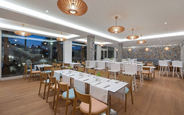 Un restaurante moderno con varias mesas y sillas, ventanas amplias y luminarias decorativas en el techo.