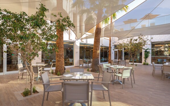 An outdoor dining area with tables and chairs, surrounded by palm trees and glass walls.