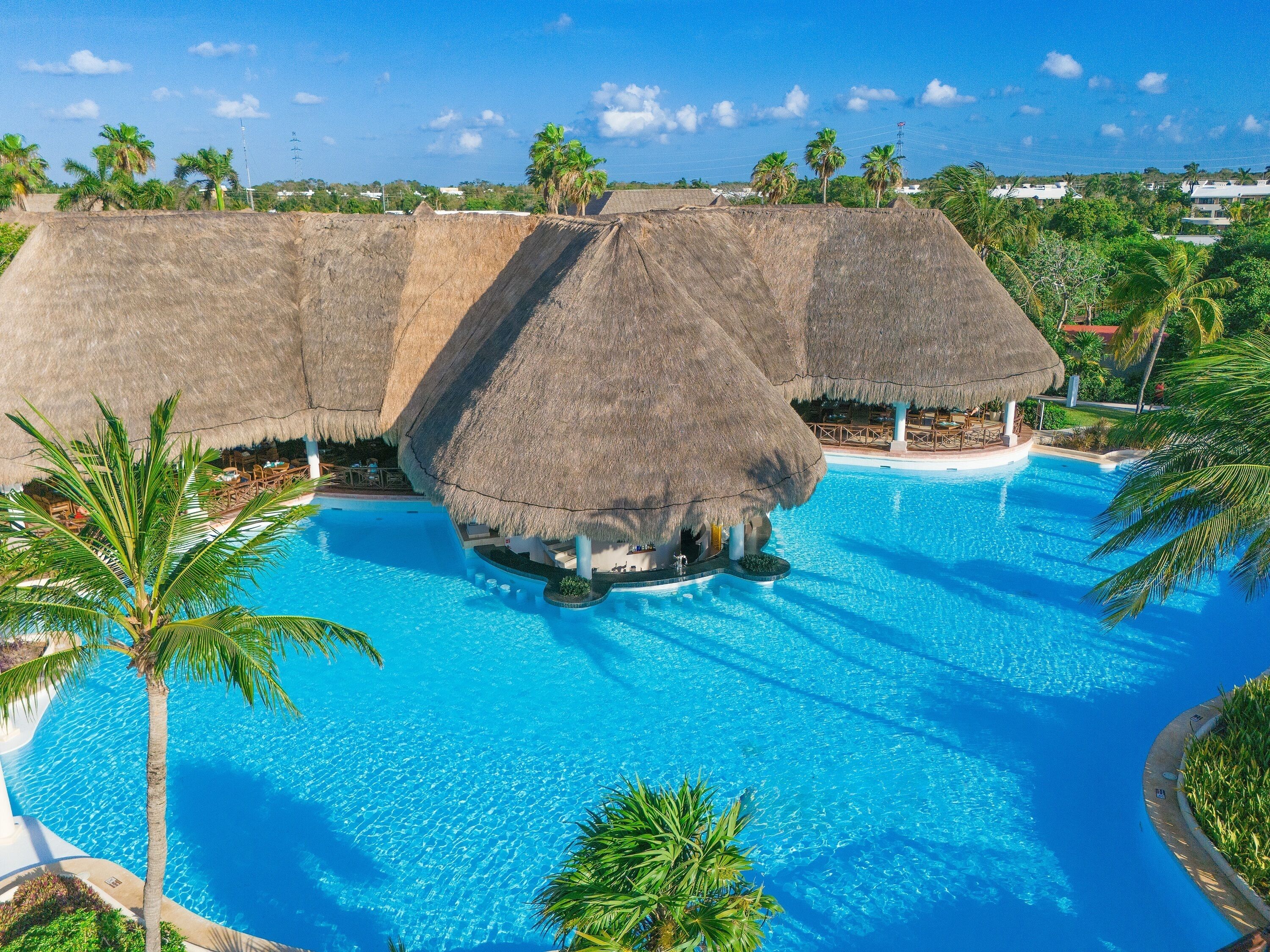 A resort with a large swimming pool, thatched-roof structures, and palm trees.