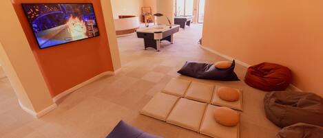 A modern living room with a flat-screen TV, a pool table, and a seating area with cushions.